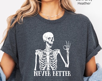 Puede incluir: Una camiseta de color gris oscuro jaspeado con un gráfico blanco de un esqueleto haciendo un gesto de "ok" con la mano. El texto "NEVER BETTER" está impreso en letras mayúsculas blancas debajo del esqueleto. La camiseta tiene mangas cortas.