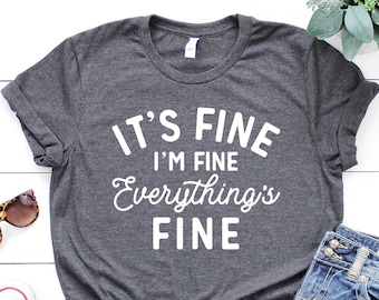 May include: A dark gray t-shirt with the text "It's Fine I'm Fine Everything's Fine" in white lettering. The shirt is laid out on a white wooden surface with a pair of sunglasses, a pair of white sneakers, and a pair of blue jeans.