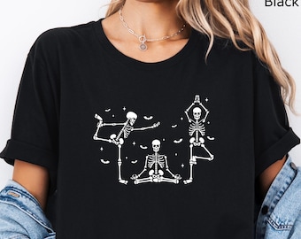 Puede incluir: Una camiseta negra de manga corta con un estampado gráfico blanco de tres esqueletos realizando posturas de yoga. Los esqueletos están rodeados de pequeñas formas blancas de murciélagos y estrellas. La camiseta tiene cuello redondo y se lleva con una chaqueta vaquera.