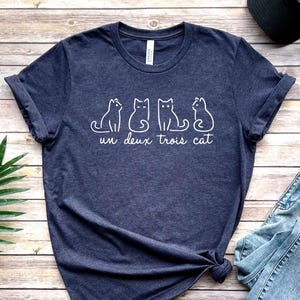 May include: A navy blue t-shirt with a white graphic of three cats and the text "un deux trois cat".