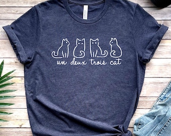 May include: A navy blue t-shirt with a white graphic of three cats and the text "un deux trois cat".