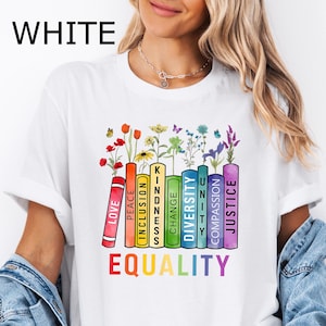 May include: White t-shirt with a rainbow-colored stack of books that have words like "Love", "Peace", "Inclusion", "Kindness", "Change", "Diversity", "Unity", "Compassion", and "Justice" printed on them. The words are arranged in a rainbow pattern. The words "EQUALITY" are printed below the books.