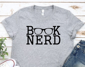 May include: A gray t-shirt with the text "BOOK NERD" and a pair of glasses printed on it. The shirt is laid out on a white wooden surface with a pair of white sneakers and blue jeans.