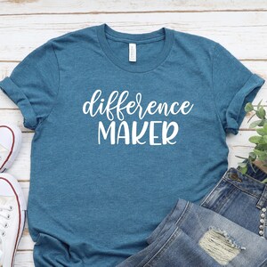 May include: A teal blue t-shirt with the text "difference maker" in white lettering. The shirt is laid out on a white wooden surface with a pair of white sneakers and a pair of blue jeans.