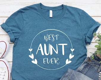 Puede incluir: Una camiseta azul turquesa con un diseño circular blanco que dice "Best Aunt Ever" en una fuente blanca manuscrita. La camiseta está colocada sobre una superficie de madera blanca con un par de zapatillas blancas y unos vaqueros azules.