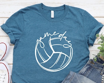 May include: Teal t-shirt with a white volleyball graphic and the text "game day". The shirt is paired with blue jeans and white sneakers. The shirt is made of a soft material.