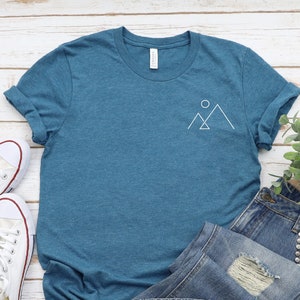 Peut inclure: Un t-shirt bleu sarcelle avec un motif blanc repr&eacute;sentant une cha&icirc;ne de montagnes et un soleil sur la poitrine gauche. Le t-shirt est pos&eacute; sur une surface en bois blanc avec une paire de baskets blanches et un jean bleu.