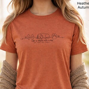 May include: A heather autumn orange short-sleeve t-shirt featuring a black line art graphic of a dog, a bone, and a paw print. Below the graphic, the text reads: Life is better with a dog OR TWO, OR THREE. The shirt has a crew neckline.