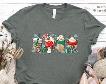 Puede incluir: Camiseta verde militar jaspeada con un diseño festivo de cuatro tazas de café temáticas navideñas y copos de nieve. Las tazas están decoradas con bastones de caramelo, crema batida y cajas de regalo. La camiseta es de un material suave.
