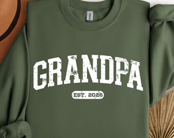 May include: Military green sweatshirt with the word "GRANDPA" in distressed white lettering. Below the word "GRANDPA" is the text "EST. 2026" in a smaller white font. The sweatshirt has a crew neck and long sleeves.