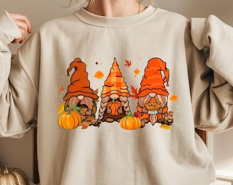 Puede incluir: Sudadera beige con un gráfico colorido de tres gnomos otoñales con sombreros naranjas, calabazas y hojas. Los gnomos tienen barbas y sostienen calabazas y tartas. La sudadera es de color neutro y tiene mangas largas.