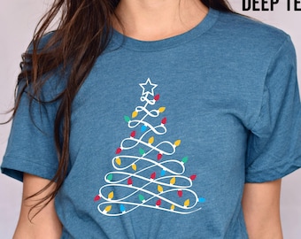 May include: A teal blue t-shirt with a white outline of a Christmas tree with colorful lights. The text "HEATHER DEEP TEAL" is in the top right corner of the image.