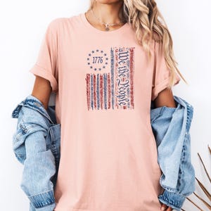 May include: Heather peach t-shirt featuring a distressed American flag design. The flag includes the text "1776" within a circle of stars and the words "We the People" in a vertical column. The shirt has rolled-up sleeves.