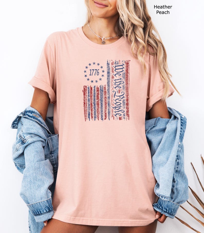 May include: Heather peach t-shirt featuring a distressed American flag design. The flag includes the text "1776" within a circle of stars and the words "We the People" in a vertical column. The shirt has rolled-up sleeves.