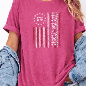May include: Heather raspberry t-shirt with a distressed American flag design. The flag features the text "1776" in a circle of stars and the words "We the People" in a vertical stripe. The shirt has rolled-up sleeves.