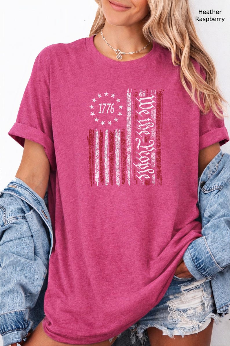 May include: Heather raspberry t-shirt with a distressed American flag design. The flag features the text "1776" in a circle of stars and the words "We the People" in a vertical stripe. The shirt has rolled-up sleeves.