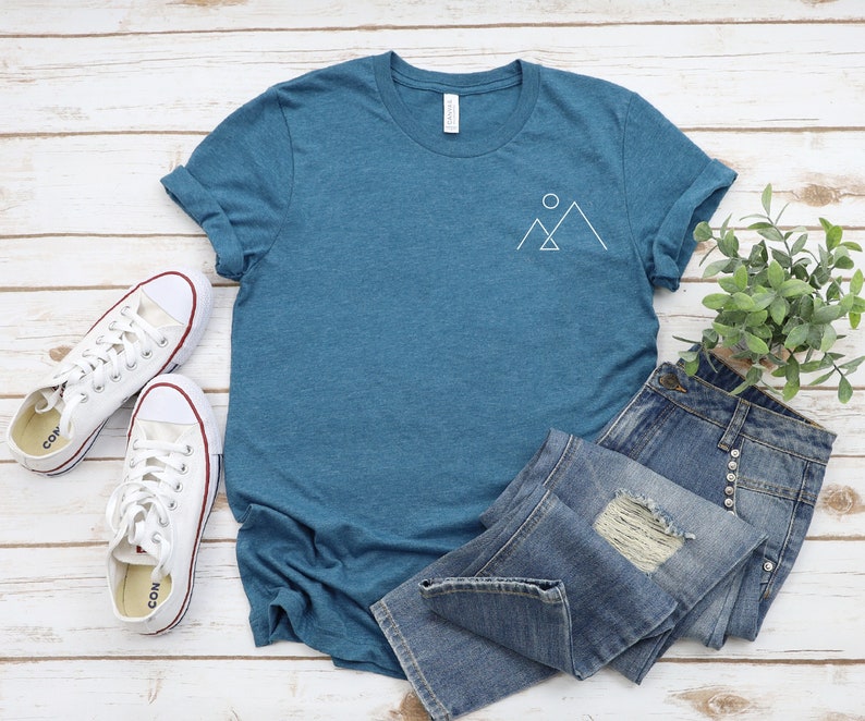 Peut inclure: Un t-shirt bleu sarcelle avec un motif blanc repr&eacute;sentant une cha&icirc;ne de montagnes et un soleil sur la poitrine gauche. Le t-shirt est pos&eacute; sur une surface en bois blanc avec une paire de baskets blanches et un jean bleu.
