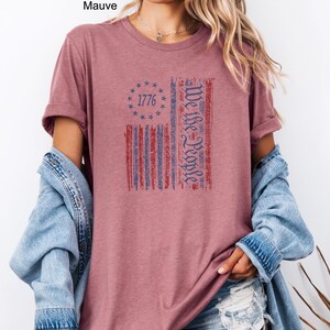 May include: Heather mauve t-shirt with a distressed American flag design. The flag features red and blue stripes, a circle of stars with the year 1776, and the text "We the People". The shirt is a solid color.