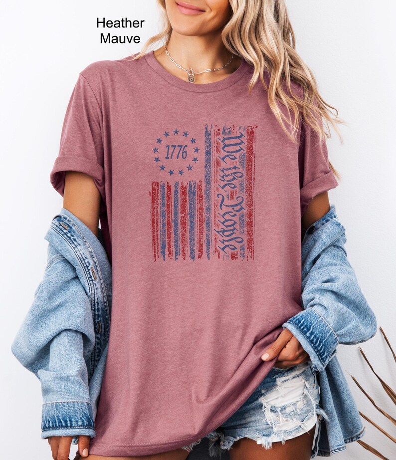 May include: Heather mauve t-shirt with a distressed American flag design. The flag features red and blue stripes, a circle of stars with the year 1776, and the text "We the People". The shirt is a solid color.