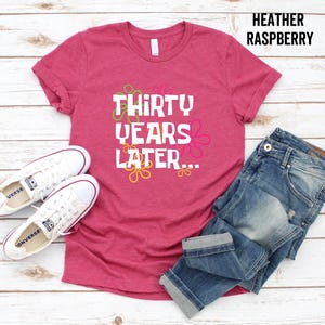 May include: A heather raspberry t-shirt with the text "Thirty Years Later..." in white with colorful flowers around the text. The shirt is shown on a wooden background with a pair of white sneakers and blue jeans.
