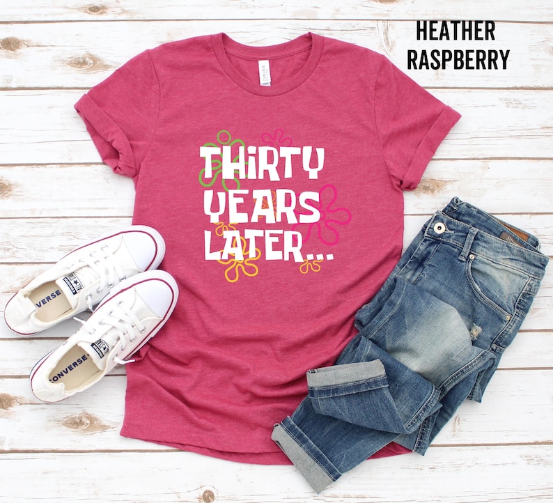 May include: A heather raspberry t-shirt with the text "Thirty Years Later..." in white with colorful flowers around the text. The shirt is shown on a wooden background with a pair of white sneakers and blue jeans.