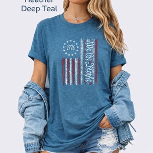 May include: Heather deep teal t-shirt with a distressed American flag design. The flag features red and white stripes, a circle of stars, and the text "1776" and "We the People" in white. The shirt has rolled-up sleeves.