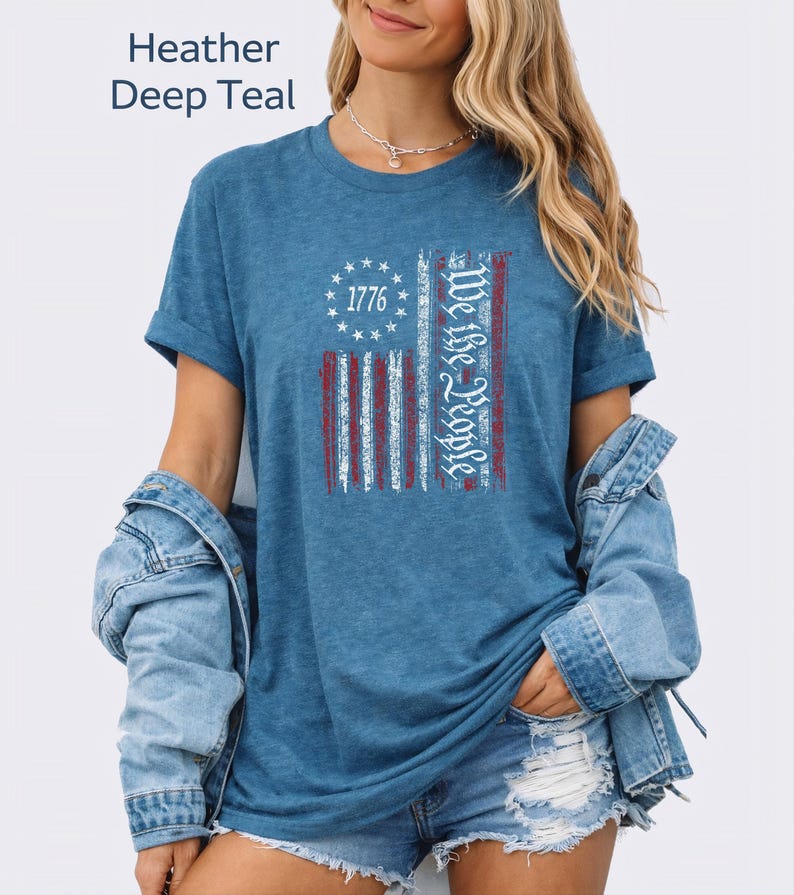 May include: Heather deep teal t-shirt with a distressed American flag design. The flag features red and white stripes, a circle of stars, and the text "1776" and "We the People" in white. The shirt has rolled-up sleeves.