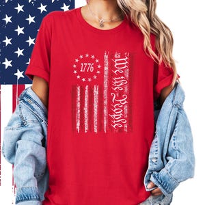 May include: Red t-shirt featuring a distressed American flag design. The flag includes the text "1776" surrounded by stars and the words "We the People" in a vertical column. The shirt has short sleeves.