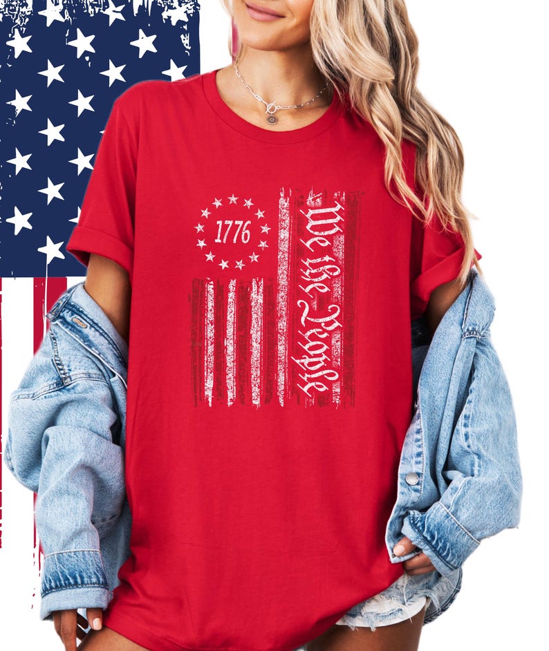 May include: Red t-shirt featuring a distressed American flag design. The flag includes the text "1776" surrounded by stars and the words "We the People" in a vertical column. The shirt has short sleeves.