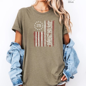 May include: Heather olive-colored t-shirt featuring a distressed American flag design. The flag includes the text "1776" in a circle of stars and "We the People" in a vertical script. The shirt has short sleeves and a relaxed fit.