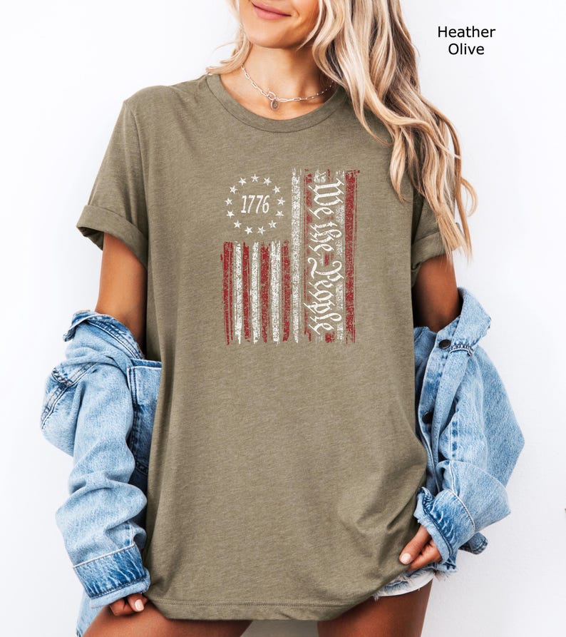 May include: Heather olive-colored t-shirt featuring a distressed American flag design. The flag includes the text "1776" in a circle of stars and "We the People" in a vertical script. The shirt has short sleeves and a relaxed fit.