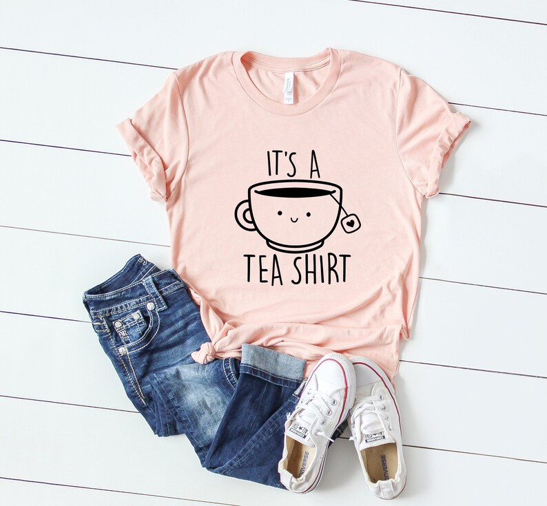 May include: A pink t-shirt with a black graphic of a teacup with a smiling face and the text "It's a Tea Shirt".