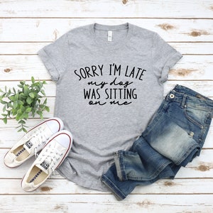 Op de afbeelding: Een grijs T-shirt met de tekst "Sorry, ik ben te laat, mijn hond zat op me" in zwarte letters. Het shirt wordt gecombineerd met een blauwe jeans en witte sneakers.