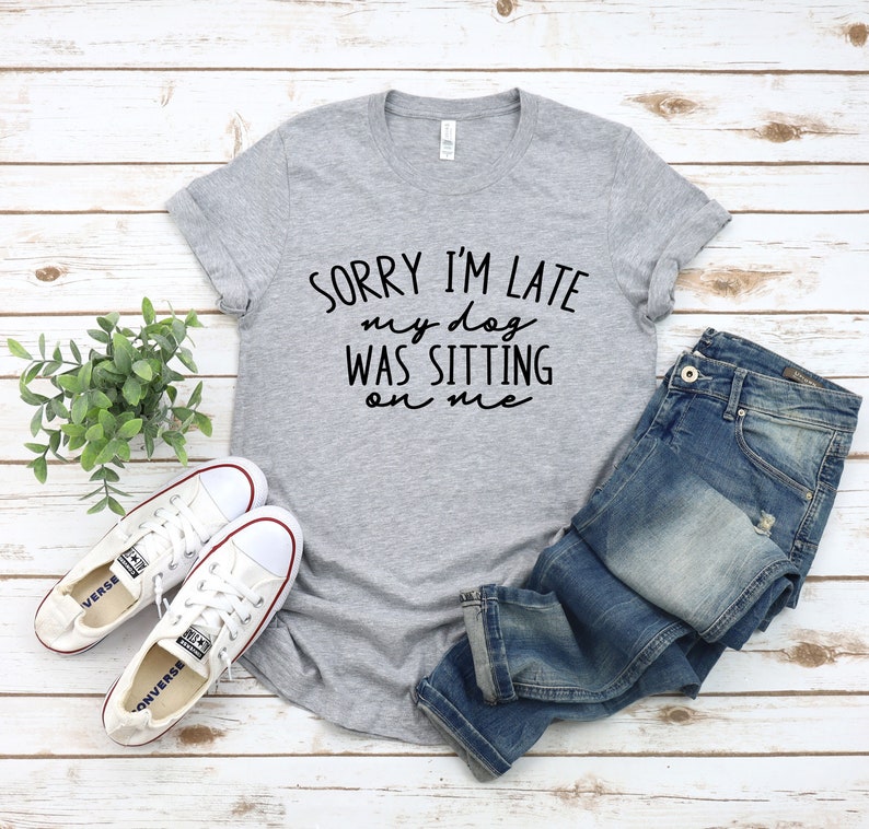 Op de afbeelding: Een grijs T-shirt met de tekst "Sorry, ik ben te laat, mijn hond zat op me" in zwarte letters. Het shirt wordt gecombineerd met een blauwe jeans en witte sneakers.