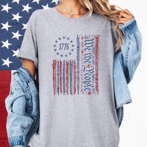 May include: A gray t-shirt featuring a distressed American flag design. The flag includes the text "1776" in a circle of stars and the words "We the People" in a vertical column. The shirt is worn with a denim jacket.