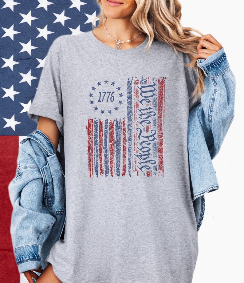 May include: A gray t-shirt featuring a distressed American flag design. The flag includes the text "1776" in a circle of stars and the words "We the People" in a vertical column. The shirt is worn with a denim jacket.