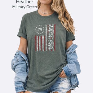 May include: Heather military green t-shirt featuring a distressed American flag design. The flag includes the text "1776" and "We the People" in white. The shirt has rolled-up sleeves.