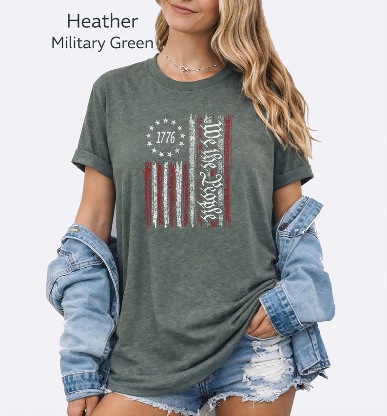 May include: Heather military green t-shirt featuring a distressed American flag design. The flag includes the text "1776" and "We the People" in white. The shirt has rolled-up sleeves.