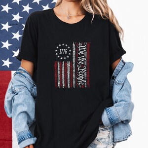 May include: Black t-shirt featuring a distressed American flag design. The flag includes the text "1776" surrounded by stars and the words "We the People" in a vertical column. The shirt is worn with a denim jacket.