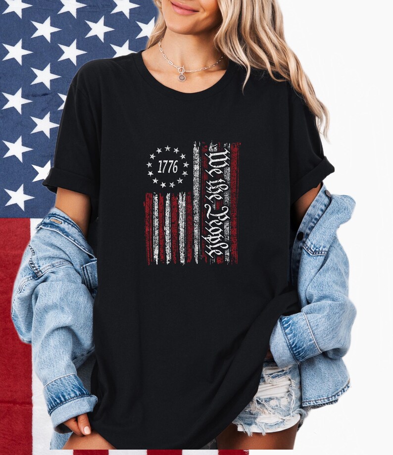 May include: Black t-shirt featuring a distressed American flag design. The flag includes the text "1776" surrounded by stars and the words "We the People" in a vertical column. The shirt is worn with a denim jacket.