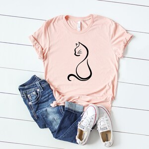 May include: A pink t-shirt with a black outline of a cat sitting on it. The shirt is laying on a white wooden surface with a pair of white sneakers and blue jeans.