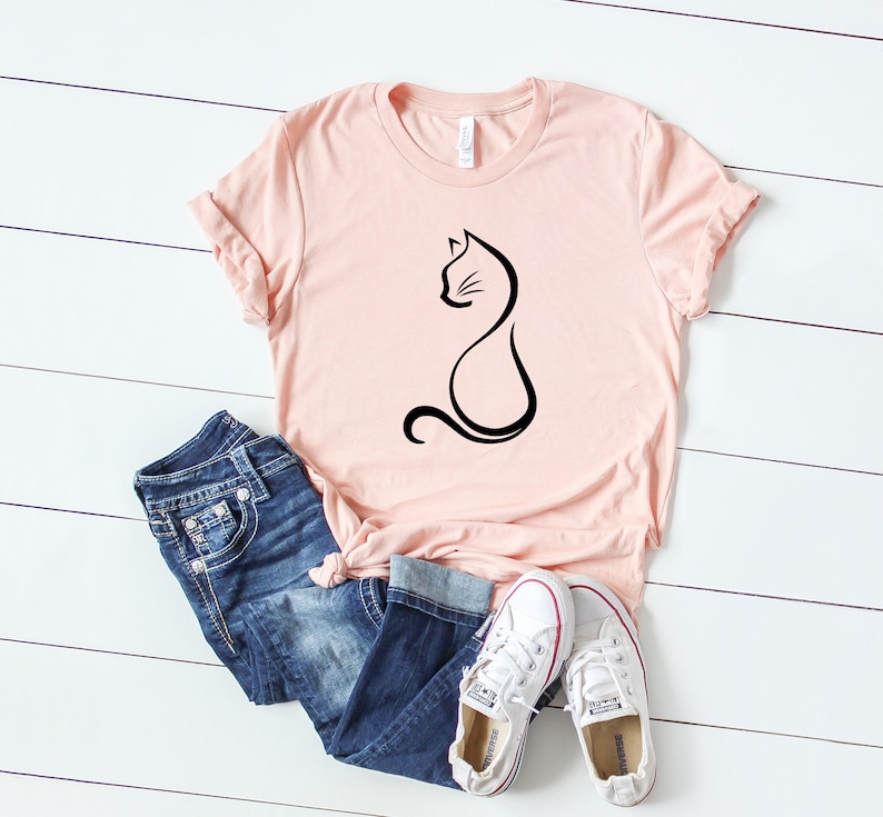 May include: A pink t-shirt with a black outline of a cat sitting on it. The shirt is laying on a white wooden surface with a pair of white sneakers and blue jeans.