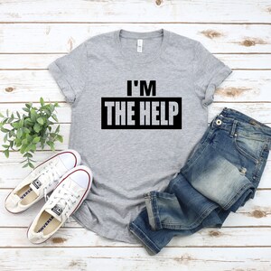 Puede incluir: Una camiseta gris con el texto "I'M THE HELP" impreso en negro en la parte delantera. La camiseta se combina con unos vaqueros azules y zapatillas blancas.