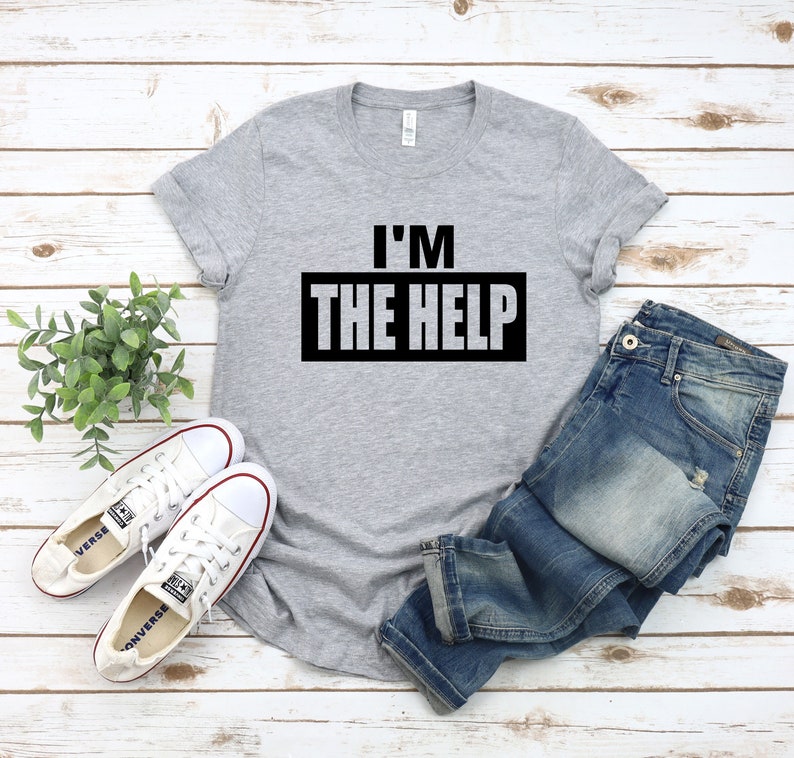 Puede incluir: Una camiseta gris con el texto "I'M THE HELP" impreso en negro en la parte delantera. La camiseta se combina con unos vaqueros azules y zapatillas blancas.