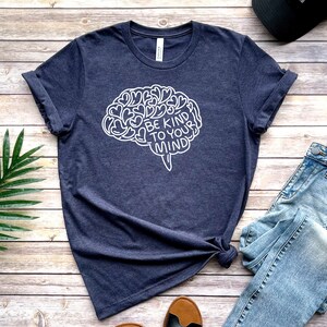 May include: A navy blue t-shirt with a white outline of a brain with the text "Be Kind To Your Mind" inside. The shirt is laid flat on a wooden surface with a pair of jeans and a hat.