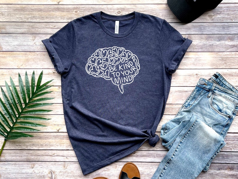May include: A navy blue t-shirt with a white outline of a brain with the text "Be Kind To Your Mind" inside. The shirt is laid flat on a wooden surface with a pair of jeans and a hat.