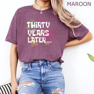 May include: A heather maroon t-shirt with the text "Thirty Years Later..." in white with colorful swirls around the text.