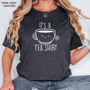 May include: A dark gray heather t-shirt with the text "It's a Tea Shirt" and a cartoon illustration of a teacup with a smiling face.