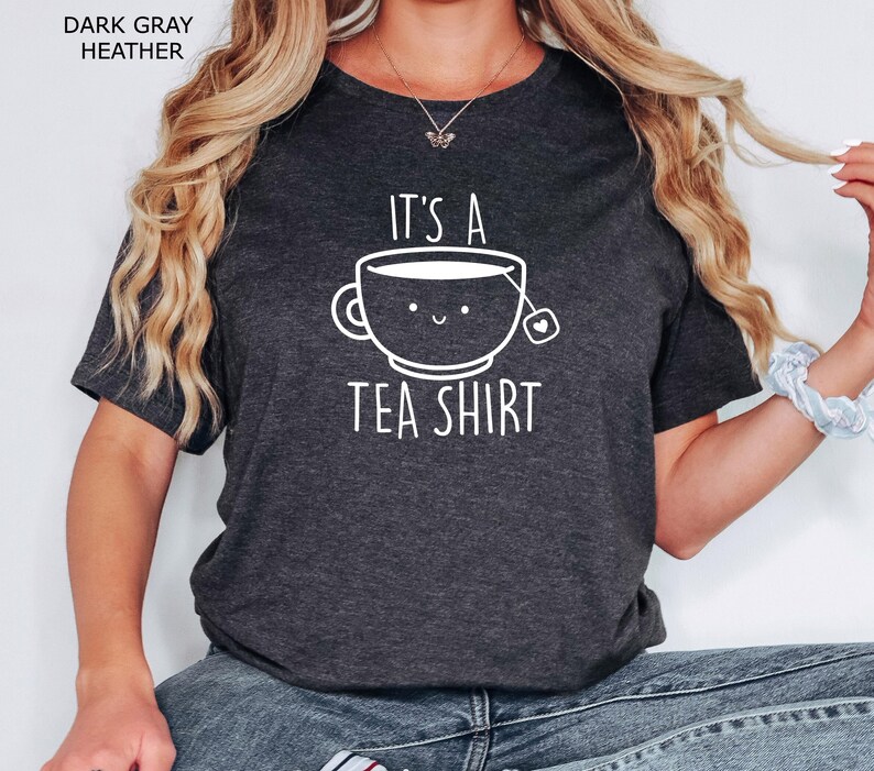 May include: A dark gray heather t-shirt with the text "It's a Tea Shirt" and a cartoon illustration of a teacup with a smiling face.