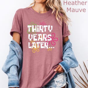 May include: Heather mauve t-shirt with a white graphic that says "Thirty Years Later..." with colorful flowers around the text.